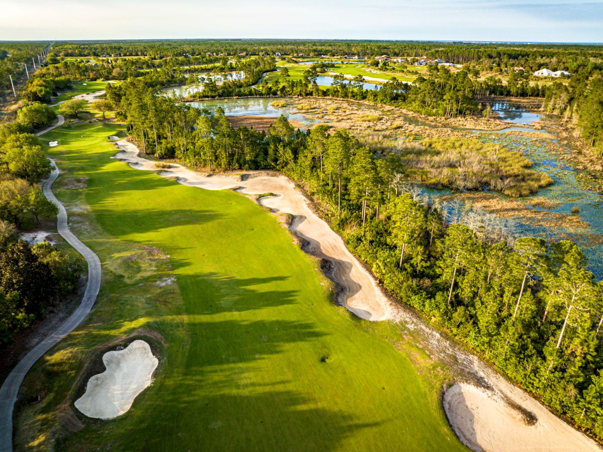 The image shows an aerial view of a golf course with sand traps, lush greenery, and a water body on the right side.