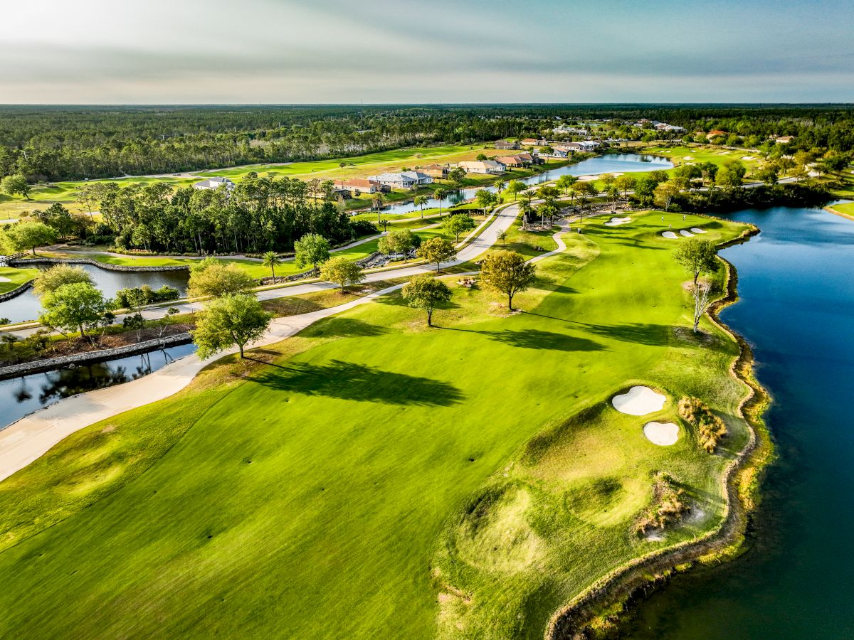 Aerial view of a lush golf course with winding paths, sand traps, and adjacent water features, surrounded by trees and residential areas.