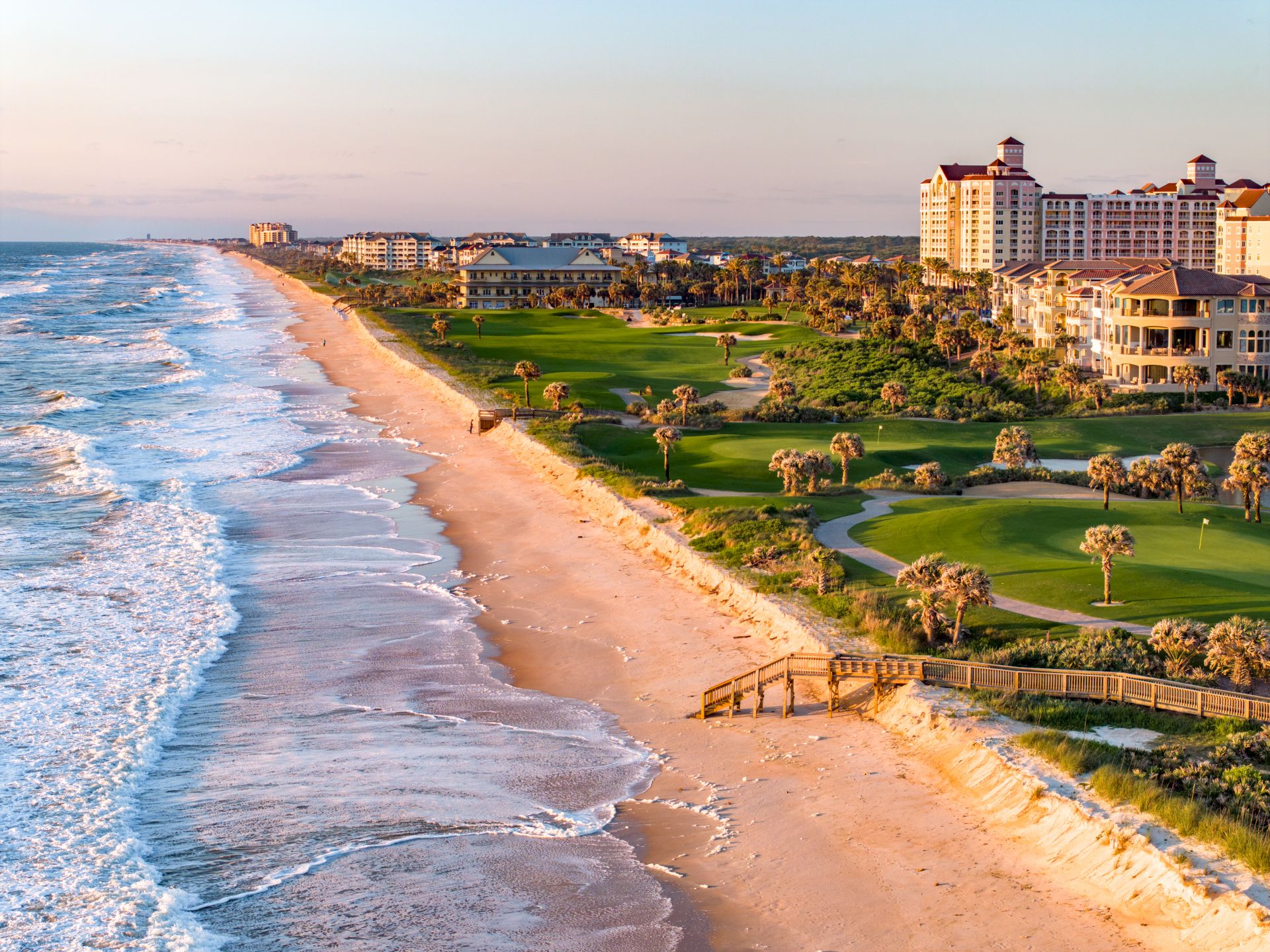 A scenic coastal view with a sandy beach, ocean waves, lush greenery, and buildings in the background during sunset.