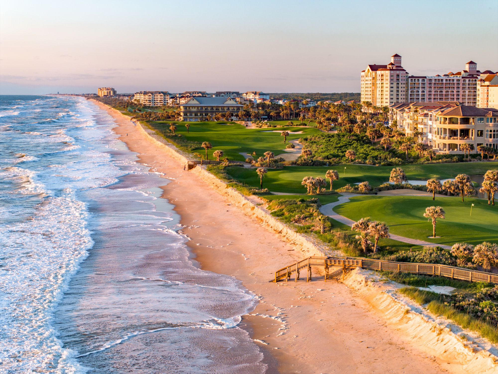 A scenic coastal view with a sandy beach, ocean waves, lush greenery, and buildings in the background during sunset.
