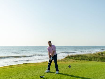 A person is playing golf near the ocean, standing on green grass with a club in hand, ready to swing.