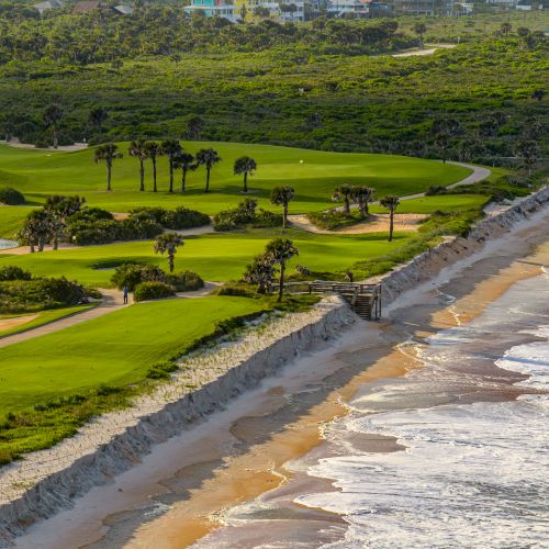 A golf course with palm trees runs alongside a sandy beach with ocean waves, bordered by a lush green landscape.