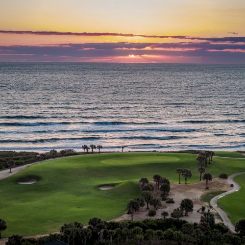 A scenic view of a golf course near the ocean at sunset with palm trees and a winding path leading to the beach in the background.