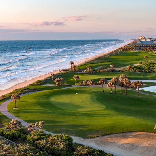 The image shows a scenic coastal golf course with green fairways, palm trees, and an ocean view at sunset.