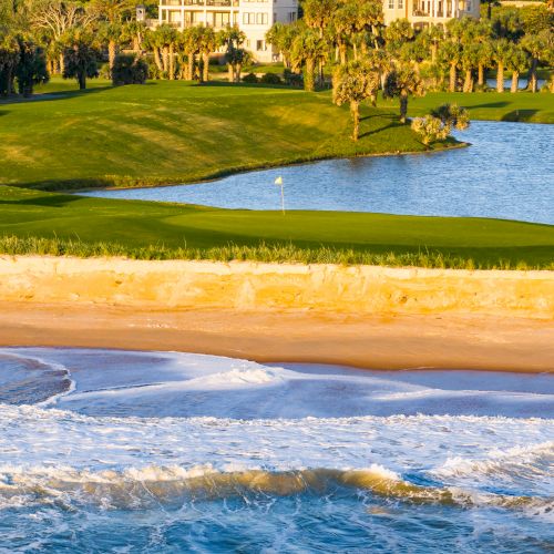 The image shows a golf course with a flag, surrounded by a body of water, palm trees, and a sandy beach with ocean waves in the foreground.