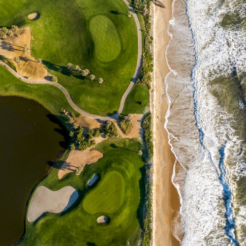An aerial view of a coastline features a beach alongside a golf course with greenery, paths, and sand traps.