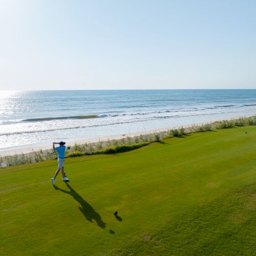 A golfer swings on a grassy course next to the ocean, under a clear blue sky with the sun reflecting on the water.