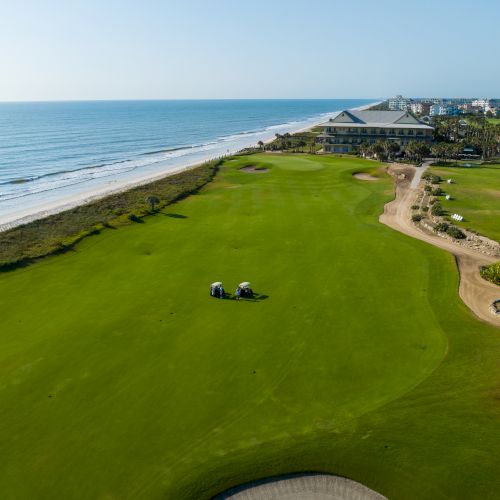 A golf course by the ocean, featuring a green, two golf carts, and a clubhouse in the distance under a clear blue sky.