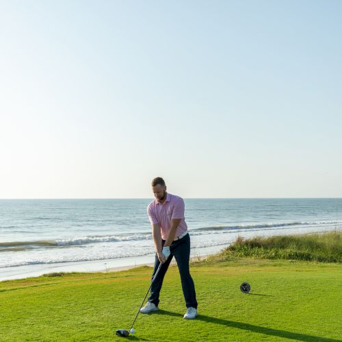 A person is playing golf on a grassy area by the ocean, preparing to swing, with the sea and sky in the background.