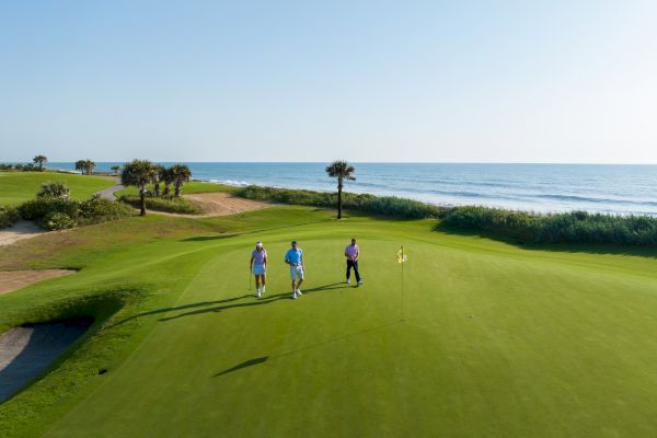 Three people walking on a golf course near the ocean, with clear skies and palm trees in the background.