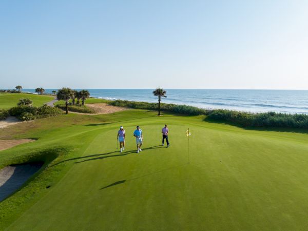 Three people walking on a golf course near the ocean, with clear skies and palm trees in the background.