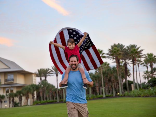 A dad carries his kids on his shoulders, wrapped in a waving American flag, with a sunny park and palm trees in the background.