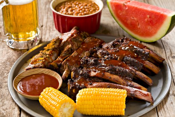 A plate with BBQ ribs, sauce, corn, baked beans; watermelon slice and beer are on the side, offering a hearty meal.