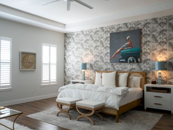 A bright bedroom with a made-up bed, patterned palm wallpaper, framed wall art, windows with blinds, and bedside lamps.