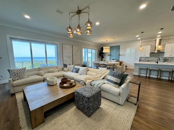 A modern living room with a sea view, featuring a large sectional sofa, wooden coffee table, and open kitchen with a dining area.