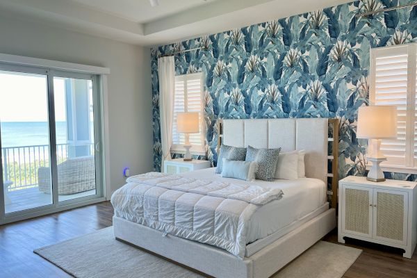 A stylish bedroom features a bed with blue pillows, a floral accent wall, nightstands with lamps, and a view of a balcony and ocean.