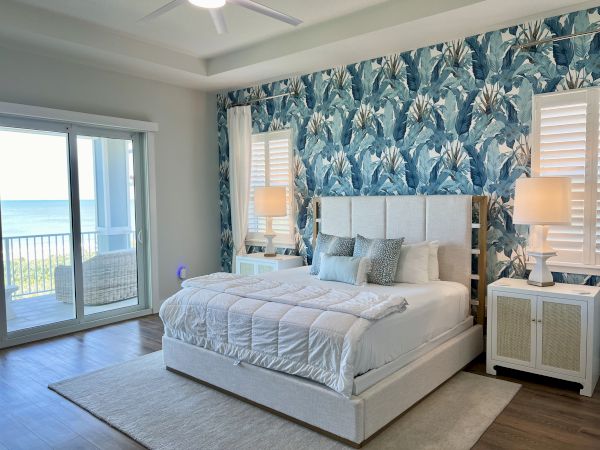 A stylish bedroom features a bed with blue pillows, a floral accent wall, nightstands with lamps, and a view of a balcony and ocean.