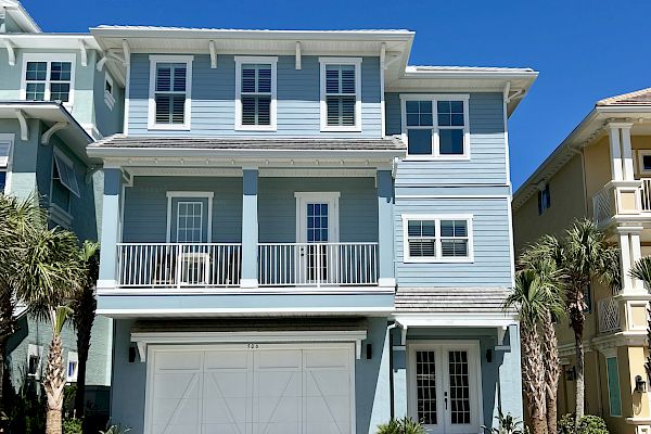 A blue, three-story house with white trim, a large garage, balconies, and palm trees in the yard under a clear blue sky.