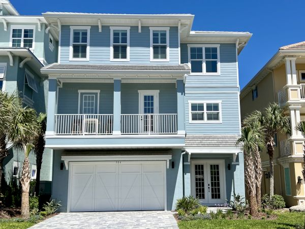 A blue, three-story house with white trim, a large garage, balconies, and palm trees in the yard under a clear blue sky.