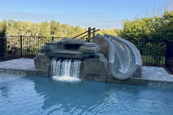 A swimming pool with a rock waterfall feature and a waterslide, surrounded by a fence and greenery in the background.