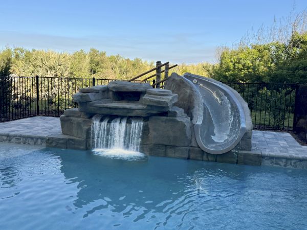 A swimming pool with a rock waterfall feature and a waterslide, surrounded by a fence and greenery in the background.