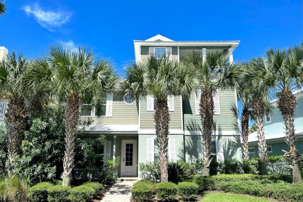The image shows a three-story house with green siding, surrounded by palm trees and neatly trimmed bushes, under a bright blue sky.