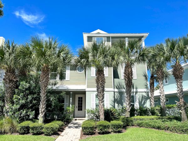 The image shows a three-story house with green siding, surrounded by palm trees and neatly trimmed bushes, under a bright blue sky.