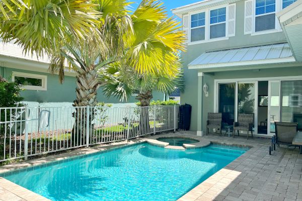 A backyard with a swimming pool, palm trees, and a two-story house with a white fence surrounding the pool area.
