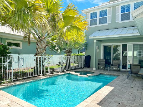 A backyard with a swimming pool, palm trees, and a two-story house with a white fence surrounding the pool area.