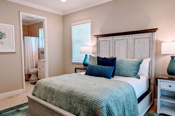 A cozy bedroom with a wooden bed, teal bedding, lamps on nightstands, and a glimpse into an adjoining bathroom with a shower.