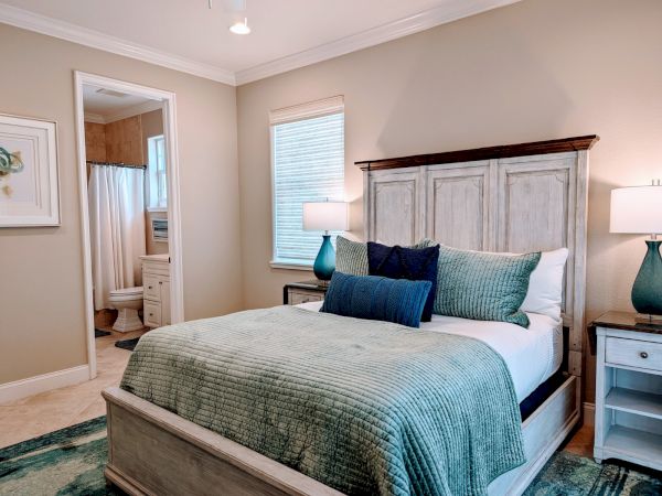 A cozy bedroom with a wooden bed, teal bedding, lamps on nightstands, and a glimpse into an adjoining bathroom with a shower.