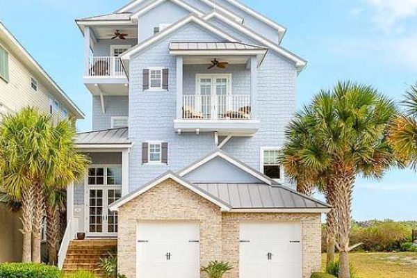A three-story, light blue beach house with balconies and palm trees in front, next to the ocean under a clear sky.