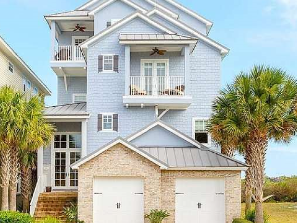 A three-story, light blue beach house with balconies and palm trees in front, next to the ocean under a clear sky.