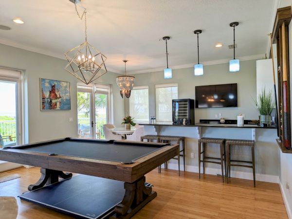 A stylish room features a pool table, a bar with stools, pendant lights, wall art, a TV, and a round dining table near large windows.