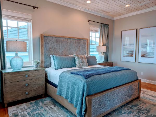 A cozy bedroom features a wooden bed with blue bedding, two nightstands with lamps, large windows, and abstract art on the walls.