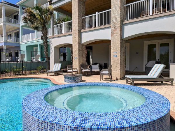 A luxurious backyard features a pool, a hot tub with blue tiles, lounge chairs, and a modern house in the background.