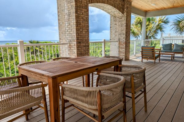 A wooden deck with a table, chairs, and a scenic view of greenery and the ocean under a cloudy sky.