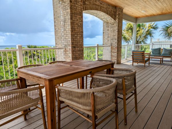 A wooden deck with a table, chairs, and a scenic view of greenery and the ocean under a cloudy sky.
