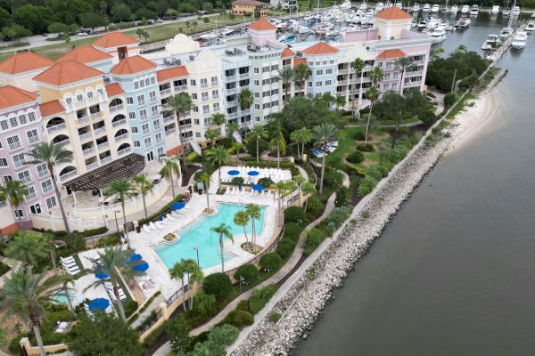 Aerial view of a waterfront resort with colorful buildings, a pool, lush gardens, and a marina with boats docked along the shore.