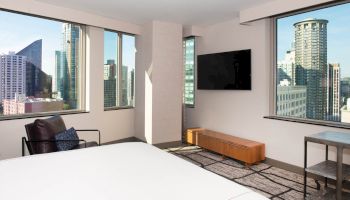 A modern hotel room with a city view, featuring a bed, a wall-mounted TV, a chair, and large windows showcasing tall buildings.