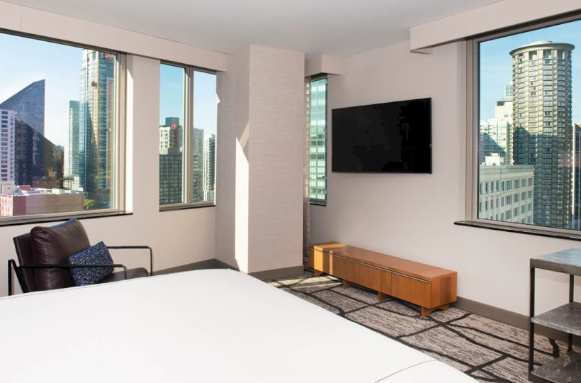 A modern hotel room with a city view, featuring a bed, a wall-mounted TV, a chair, and large windows showcasing tall buildings.