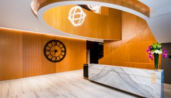 A modern lobby with a curved wooden ceiling, a large wall clock, a marble reception desk, and decorative lighting and flowers.