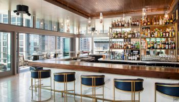A modern bar with blue stools, a wooden counter, and shelves stocked with bottles in a well-lit room with large windows overlooking a cityscape.