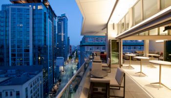 A modern cityscape view from a terrace, with tall buildings, outdoor seating, and evening lighting creating an inviting atmosphere.