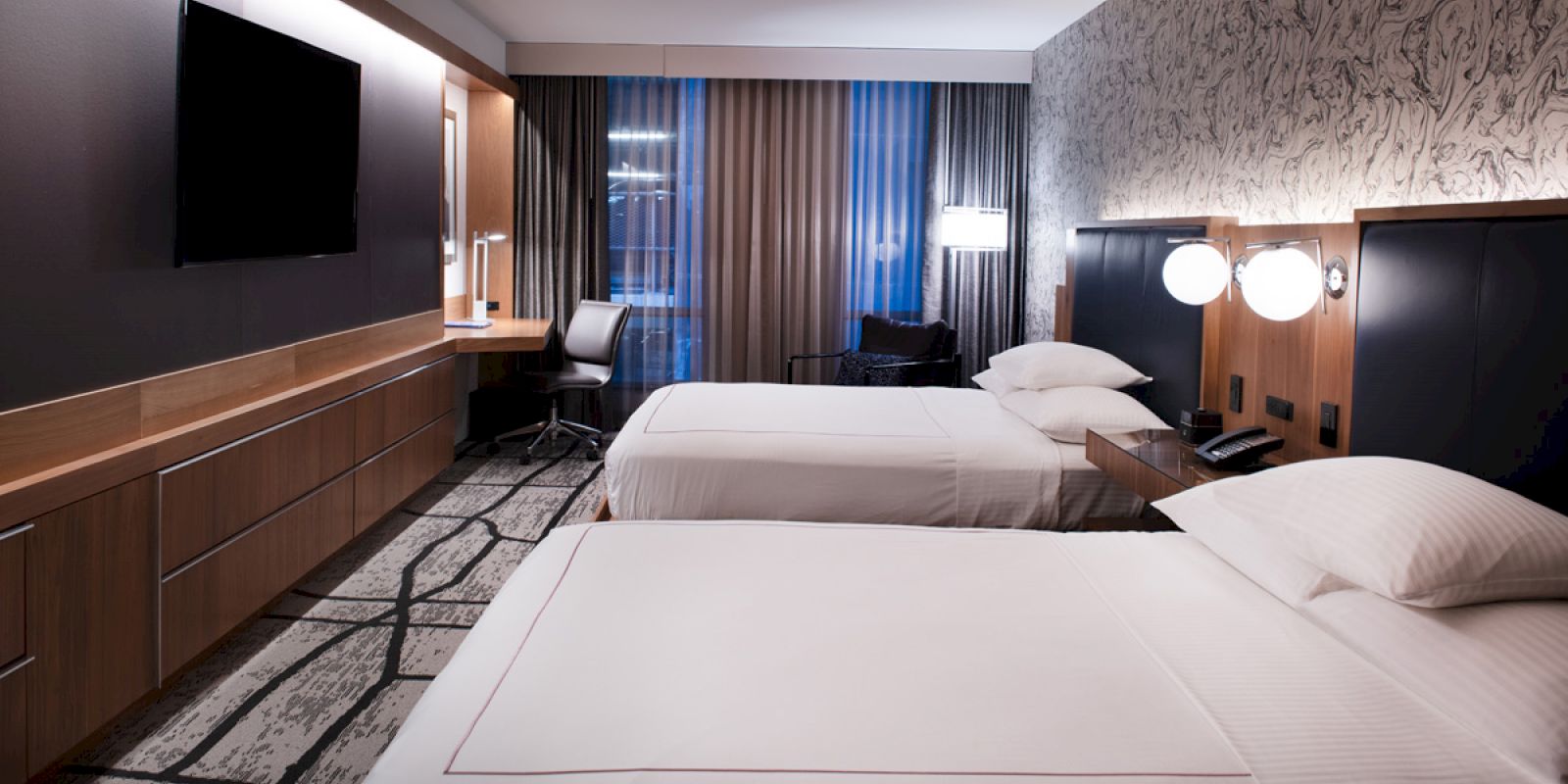 The image shows a modern hotel room with two beds, a flat-screen TV, a desk with a chair, and stylish lighting and decor.