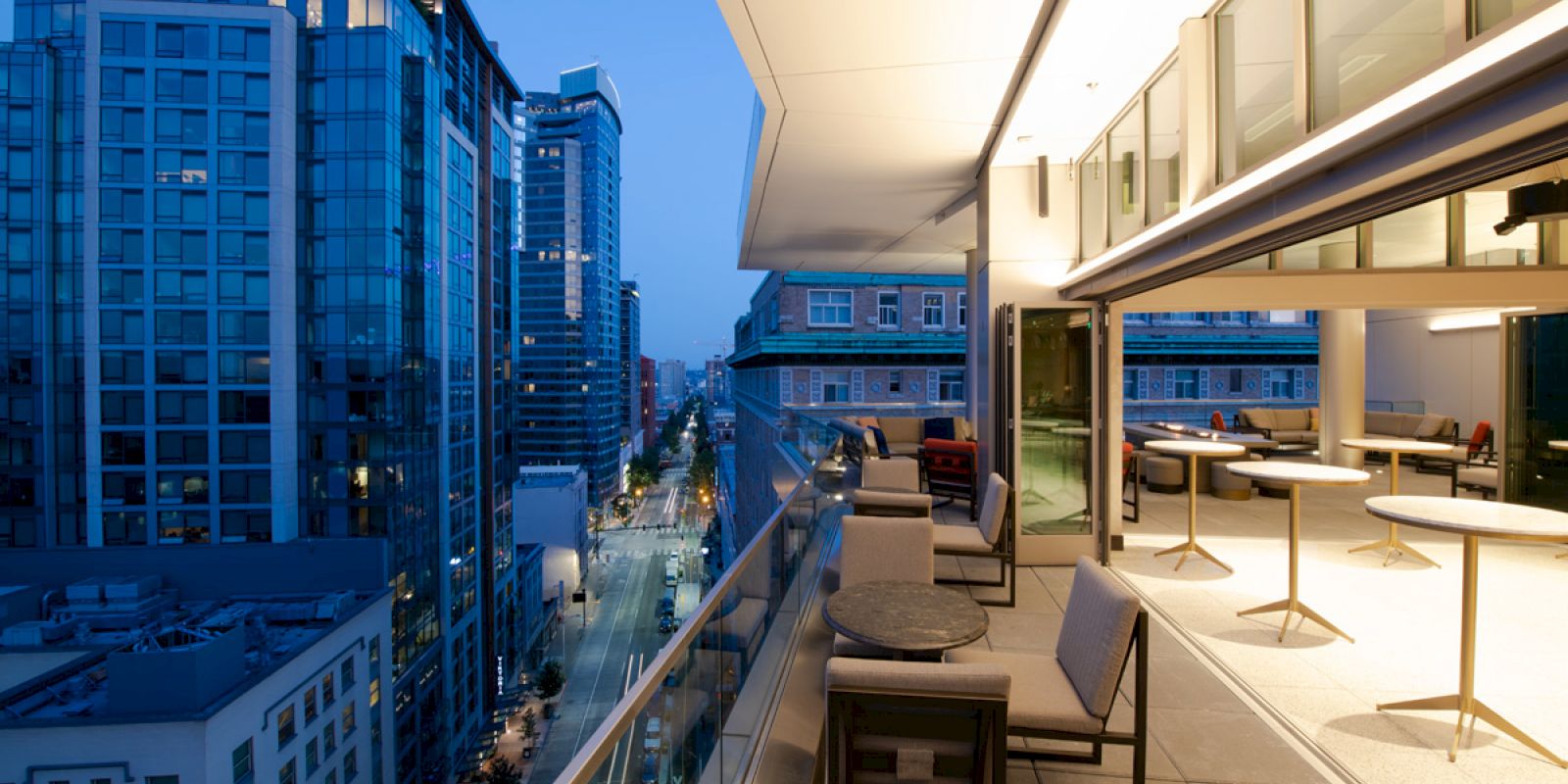 A modern cityscape at dusk with tall buildings and a furnished balcony overlooking a street.