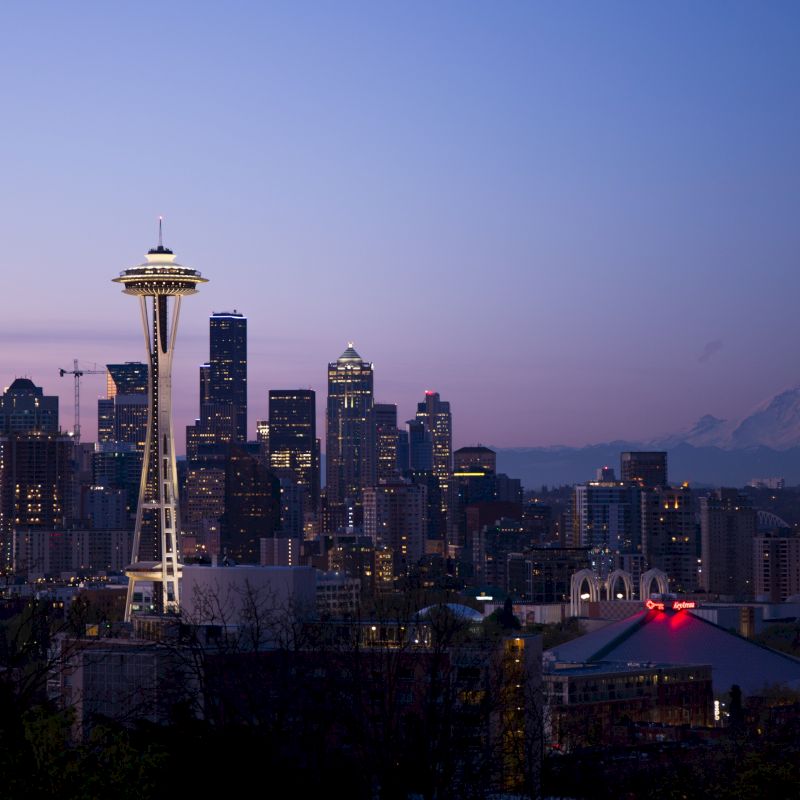 Seattle skyline at dusk, featuring the Space Needle and buildings, with Mount Rainier in the background under a clear sky.