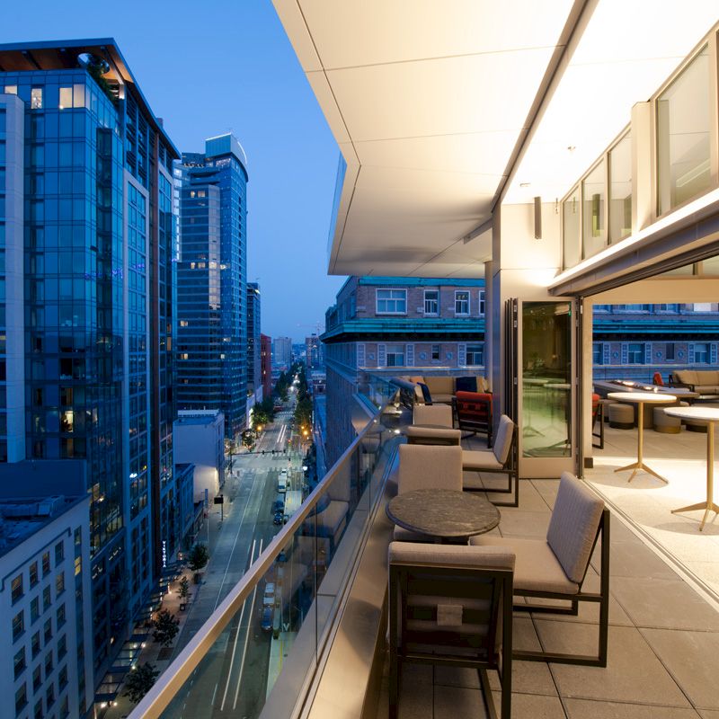 A modern outdoor balcony with seating overlooks a cityscape at dusk, with buildings lit up and a clear sky.