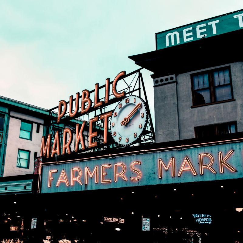 The image shows a public market with a neon "Farmers Market" sign and a clock on a building facade, evoking a busy urban scene.