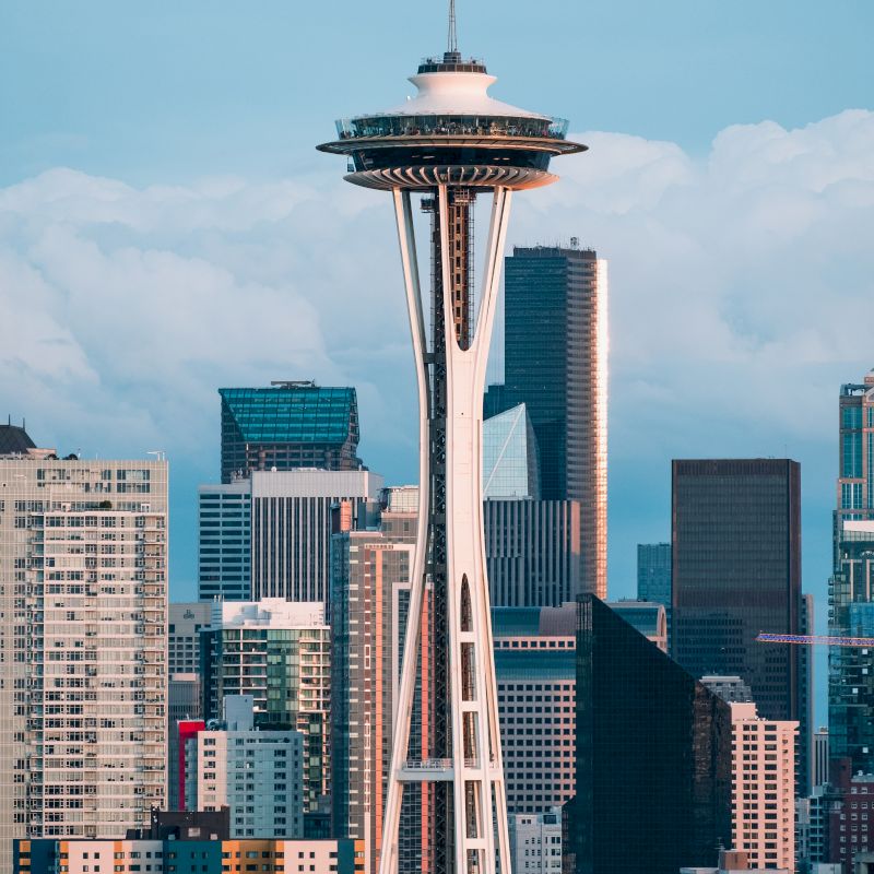 The image shows the Space Needle with a backdrop of Seattle's downtown skyline and various skyscrapers under a cloudy sky.